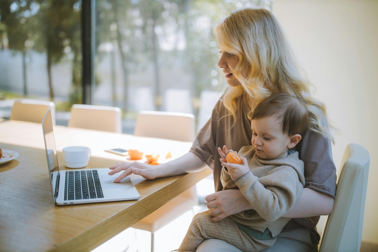 A mother holding her baby while working at a laptop