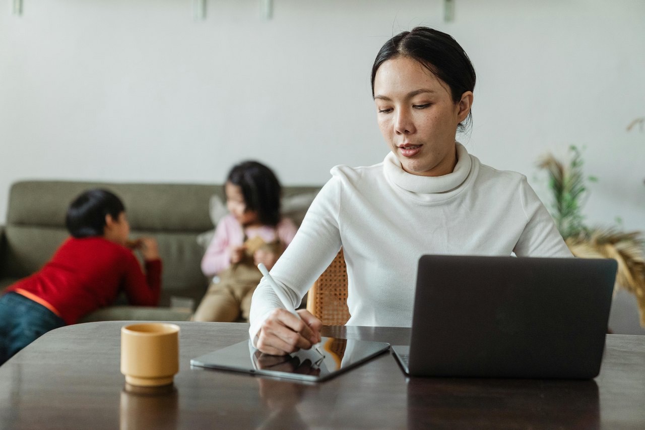 A mother working at a laptop with her young children beside her